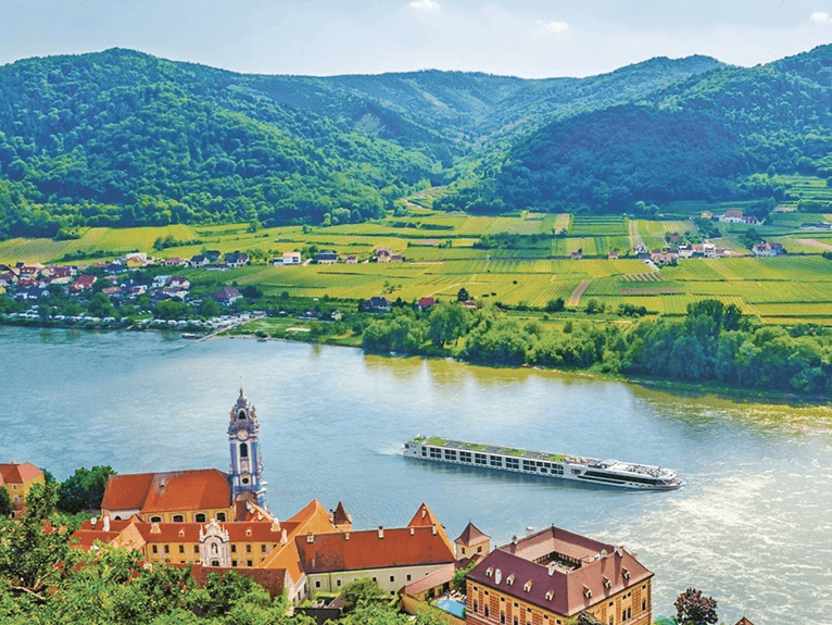 A Scenic Space-Ship cruising the Danube River next to a village and green mountains in Durnstein, Austria