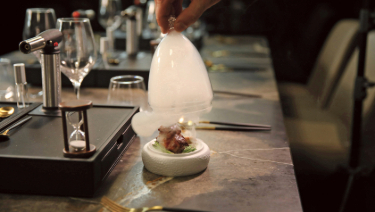 The waiter lifts a smoke filled glass above a dish, showcasing the prepared meal in a focused manner.