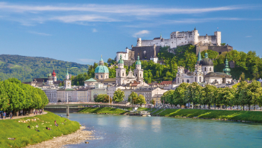 A picturesque river flowing beneath a majestic castle perched on a hill, surrounded by lush greenery and clear blue skies.