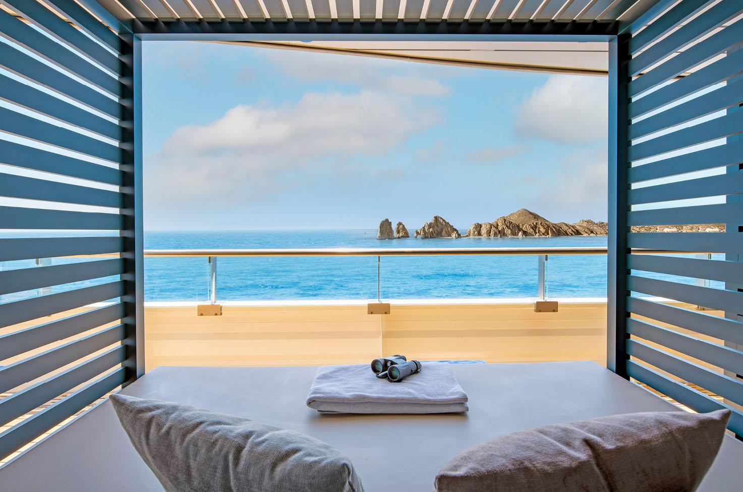 View from a cabana sunbed with cushions, towels and binoculars on board Scenic Eclipse, looking out on to the blue ocean and some grey rocky coastline
