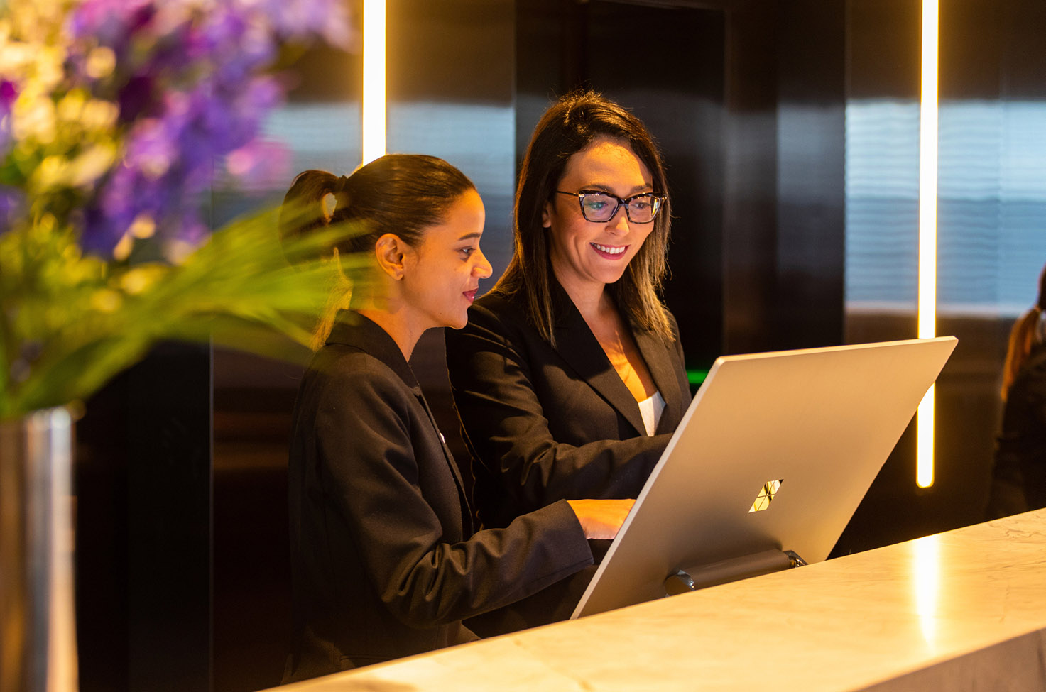 Two crew members at the reception of Scenic Eclipse looking at a screen with purple flowers in the foreground 