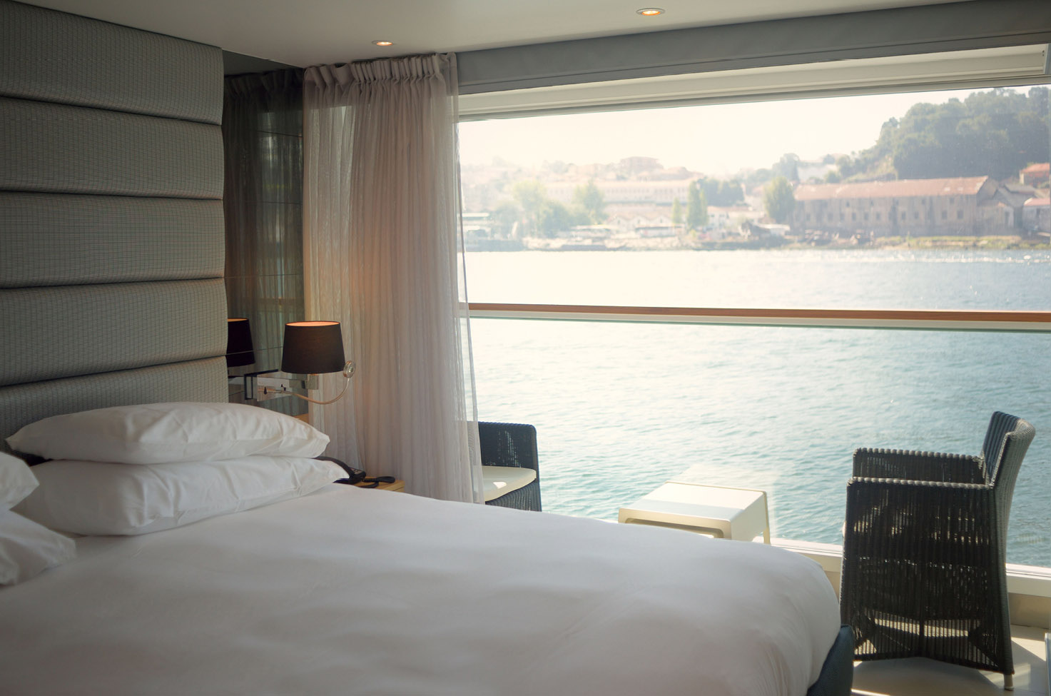 View from the window of a balcony suite with a bed and table and chair, with the banks of the Douro river outside.