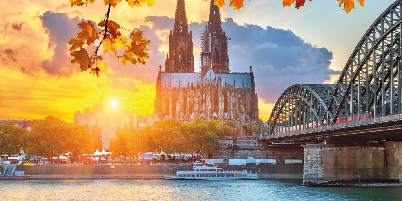 A scenic bridge spans over water with a castle visible in the background, surrounded by trees and clouds. The scene captures the essence of autumn during sunset, with reflections on the river.