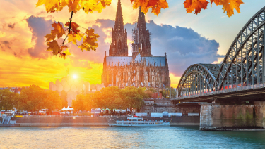 A scenic bridge spans over water with a castle visible in the background, surrounded by trees and clouds. The scene captures the essence of autumn during sunset, with reflections on the river.