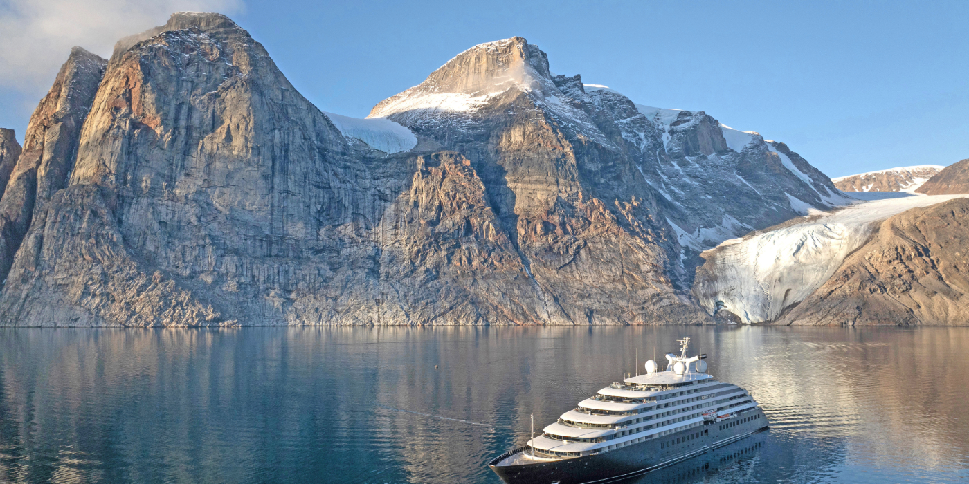 A ship sailing on a glacial lake surrounded by snow-capped mountains, with a clear blue sky above and a tranquil landscape in the background.