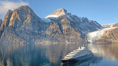 A ship sailing on a glacial lake surrounded by snow-capped mountains, with a clear blue sky above and a tranquil landscape in the background.