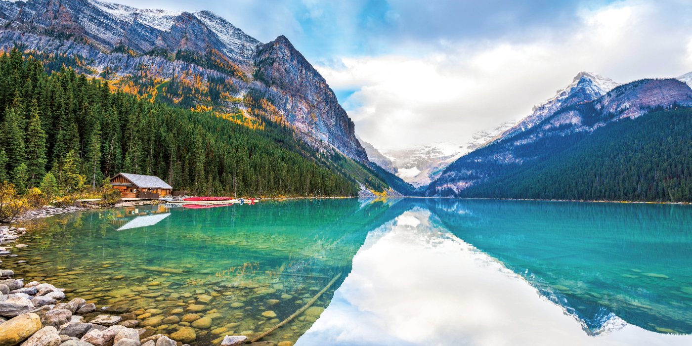 A serene body of water surrounded by mountains and trees, with a clear sky and clouds reflecting on the calm surface of the glacial lake.
