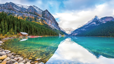 A serene body of water surrounded by mountains and trees, with a clear sky and clouds reflecting on the calm surface of the glacial lake.