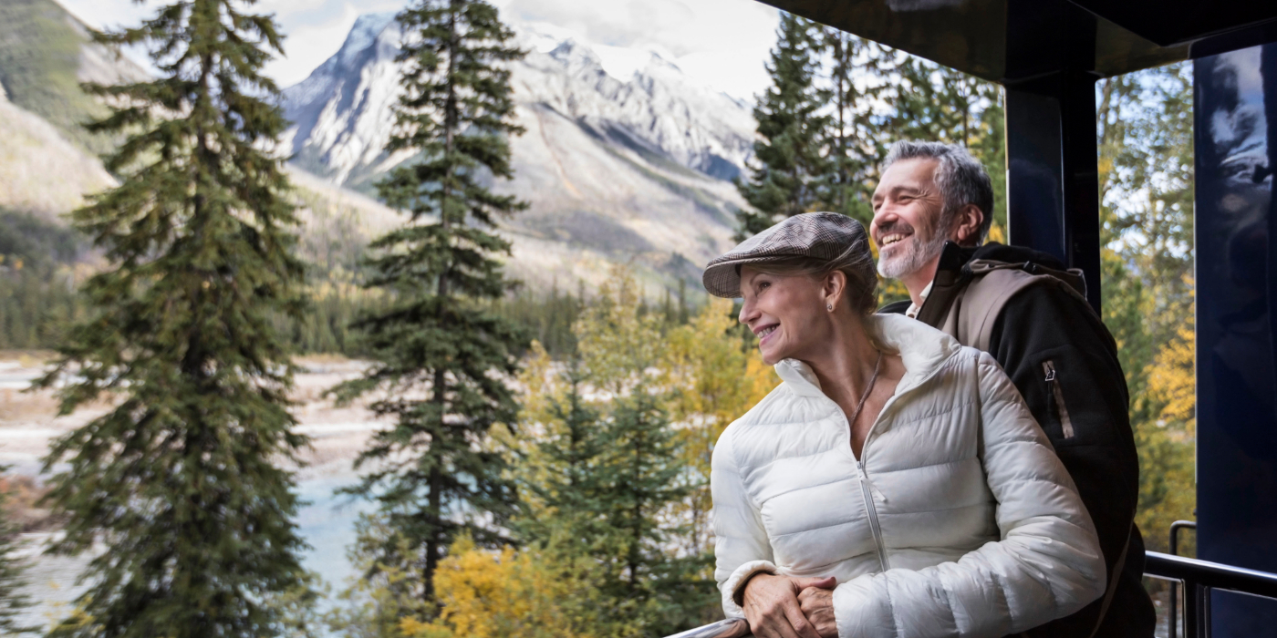 A man and a woman standing on a porch, looking at mountains in the distance. They are surrounded by trees, with the outdoor scenery enhancing the view. The man and woman are dressed casually and appear to be enjoying the moment together.