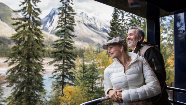A man and a woman standing on a porch, looking at mountains in the distance. They are surrounded by trees, with the outdoor scenery enhancing the view. The man and woman are dressed casually and appear to be enjoying the moment together.