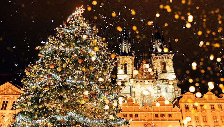 A christmas tree covered with lights, baubles and decorations in front of an illuminated building in Prague.